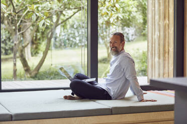 Smiling bearded mature man using laptop while sitting on bed by window at home - MCF01380