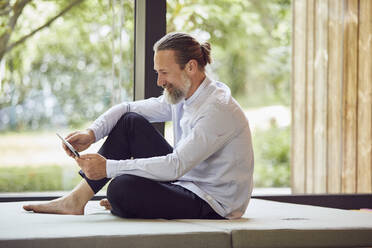 Smiling bearded mature man using digital tablet while sitting on bed by window at home - MCF01381