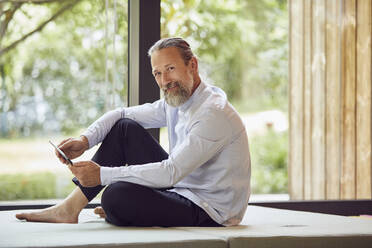 Smiling bearded man using digital tablet while relaxing on bed by window at home - MCF01382