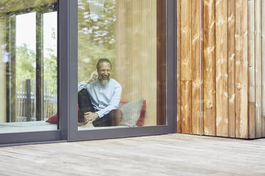 Cheerful man talking over smart phone while sitting on bed in tiny house seen through window - MCF01384
