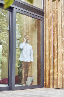 Älterer Mann, der über ein Smartphone spricht, während er in einem kleinen Haus steht, das durch ein Fenster gesehen wird - MCF01385