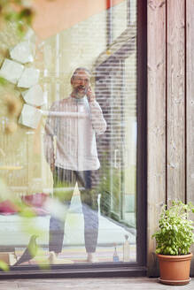 Man talking over mobile phone while standing in tiny house seen through window - MCF01386