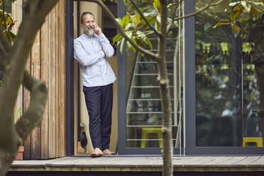 Bearded mature man talking over smart phone while standing at entrance of in tiny house - MCF01389