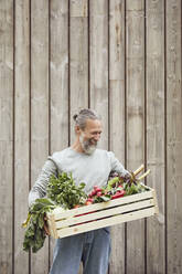 Bearded mature man carrying vegetable crate while standing against wall - MCF01396