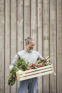 Bärtiger reifer Mann, der eine Gemüsekiste trägt, während er an der Wand steht - MCF01396
