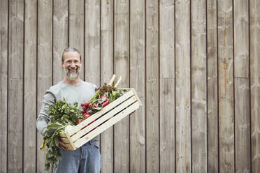 Smiling mature man carrying vegetable crate while standing against house - MCF01397