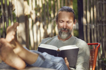 Bärtiger reifer Mann liest ein Buch und entspannt sich auf einem Stuhl im Garten - MCF01401