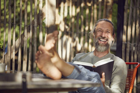 Cheerful bearded man reading book while relaxing on chair in yard - MCF01402