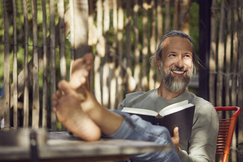 Lächelnder bärtiger Mann liest ein Buch und entspannt sich auf einem Stuhl im Garten, lizenzfreies Stockfoto