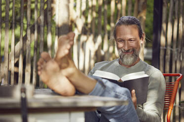 Lächelnder bärtiger Mann liest ein Buch und entspannt sich auf einem Stuhl im Garten - MCF01404