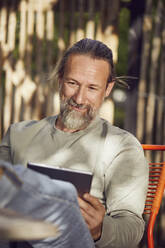 Nahaufnahme eines bärtigen Mannes, der ein Buch liest und sich auf einem Stuhl im Garten ausruht - MCF01405