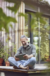 Fröhlicher bärtiger Mann, der ein digitales Tablet benutzt, während er vor einem kleinen Haus sitzt - MCF01420