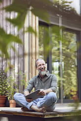 Lächelnder bärtiger Mann mit digitalem Tablet, der wegschaut, während er vor einem kleinen Haus sitzt - MCF01421