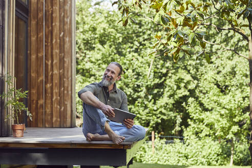 Smiling man with digital tablet looking away while sitting outside tiny house - MCF01422