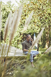 Mature man talking over smart phone while sitting against trees in yard - MCF01424