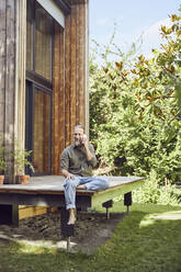 Bearded mature man talking over mobile phone while sitting outside tiny house - MCF01425