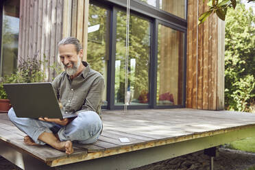 Bearded mature man using laptop while sitting outside tiny house - MCF01426