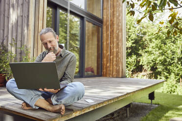 Bearded man using laptop while sitting outside house - MCF01427