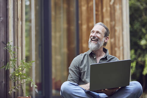 Cheerful mature man with laptop looking away while sitting outside tiny house - MCF01430