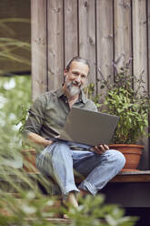 Lächelnder reifer Mann mit Laptop, der an einem kleinen Haus sitzt - MCF01432
