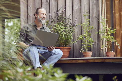 Thoughtful bearded man with laptop sitting outside tiny house - MCF01433