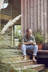 Thoughtful mature man holding coffee cup while sitting outside tiny house - MCF01434
