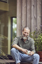 Cheerful mature man holding coffee cup while sitting outside tiny house - MCF01435