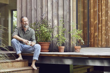 Smiling bearded man holding coffee cup while sitting outside tiny house - MCF01436