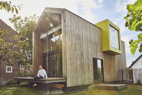 Mature man sitting outside tiny house in yard during sunny day - MCF01443