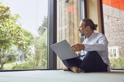 Bearded mature man with laptop looking through window while sitting in tiny house - MCF01445