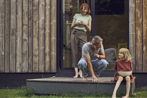 Woman looking at father and daughter eating cherries outside tiny house - MCF01449