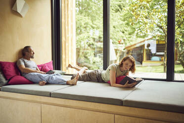 Eine Frau liest ein Buch, während ein Mann mit einem digitalen Tablet auf dem Bett am Fenster sitzt - MCF01454