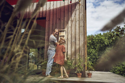 Smiling father and daughter looking away while installing insect hotel on wall - MCF01459