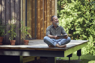 Bearded mature man holding digital tablet contemplating while sitting outside tiny house - MCF01465