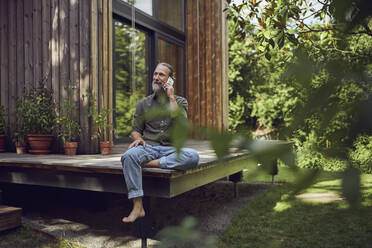 Mature man looking away while talking over mobile phone outside tiny house - MCF01467