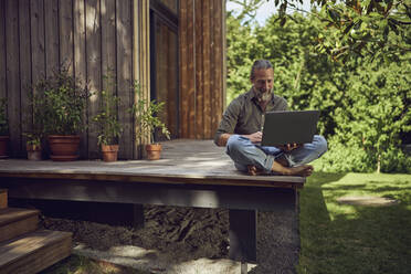 Älterer Mann, der einen Laptop benutzt, während er an einem kleinen Haus sitzt - MCF01468