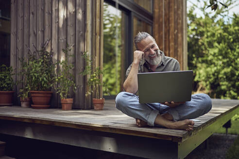 Smiling mature man using laptop while sitting against tiny house - MCF01469