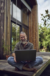 Lächelnder reifer Mann mit Laptop, der an einem kleinen Haus sitzt - MCF01470