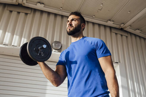 Sportsman lifting dumbbell while standing against wall - MIMFF00192