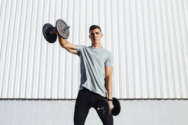 Athlete exercising with dumbbell while standing against wall - MIMFF00195