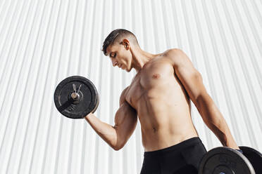 Shirtless athlete lifting dumbbell while standing against wall - MIMFF00198