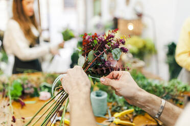 Florist bindet Blumensträuße mit der Hand, während im Hintergrund ein Team im Blumenladen arbeitet - MRRF00399