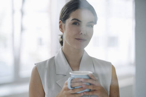 Woman holding coffee cup while standing at office - KNSF08427