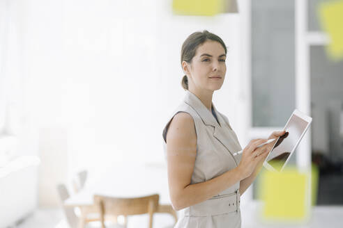 Selbstbewusste Geschäftsfrau, die im Stehen im Büro ein digitales Tablet benutzt - KNSF08446
