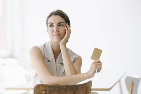 Young woman holding business card while sitting on chair at office - KNSF08459