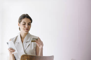 Smiling businesswoman holding credit card while using mobile phone at office - KNSF08461