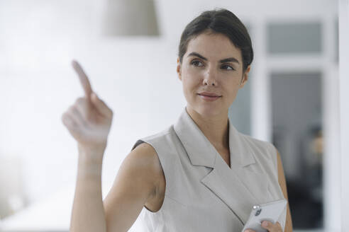 Businesswoman pointing at glass wall while standing at office - KNSF08470