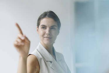 Smiling woman pointing on glass wall while standing at office - KNSF08492