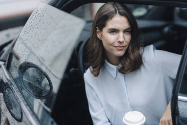 Young woman holding coffee cup while sitting in car - KNSF08519