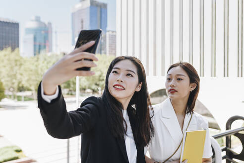 Female colleagues taking selfie on smart phone against financial district in city during sunny day - MRRF00431